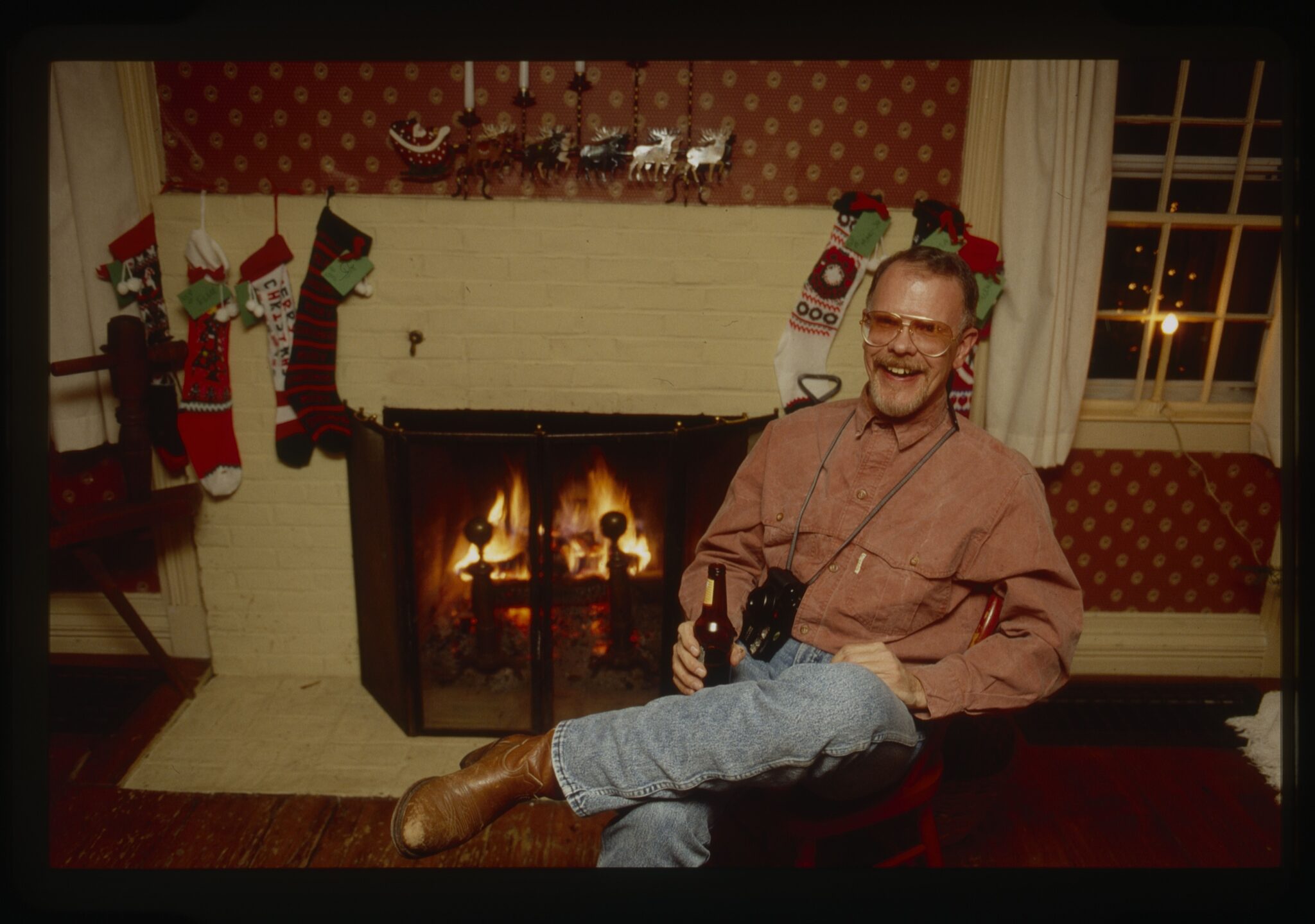 Larry Griffin by the fireplace at Christmas, camera around his neck, in jeans and cowboy boots